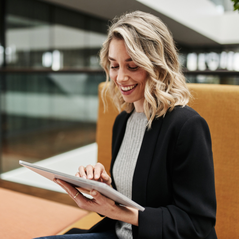 Frau sitzt auf Sofasessel und blickt lächelnd in ihr Tablet, das sie in den Händen hält