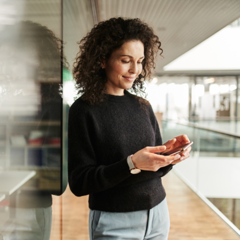 Frau lehnt an Glasfront und blickt lächelnd auf ihr Smartphone