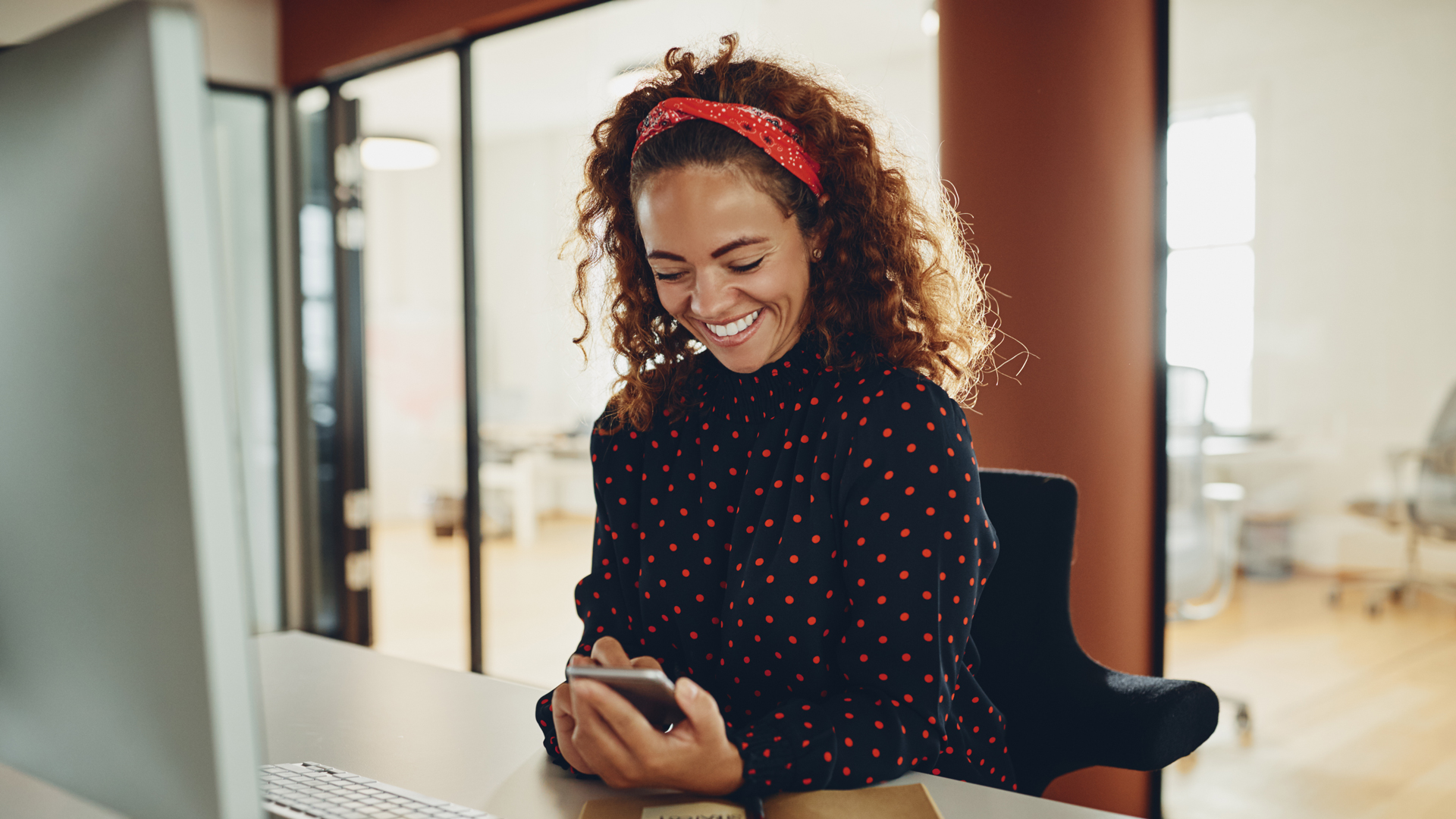 Frau sitzt am Schreibtisch und blickt lächelnd auf ihr Smartphone